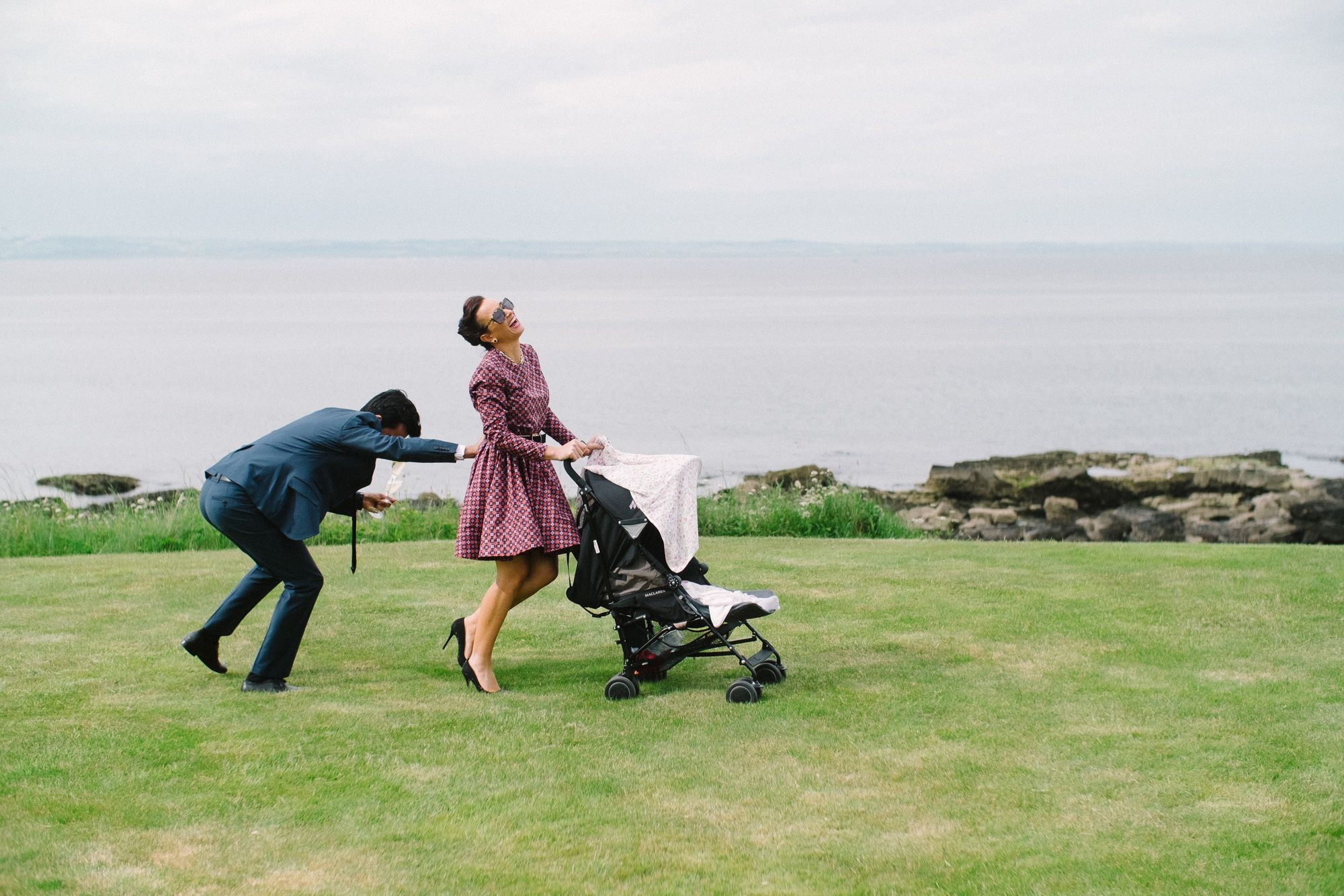 Husband helps wife pushing pram on the grass at seaside wedding